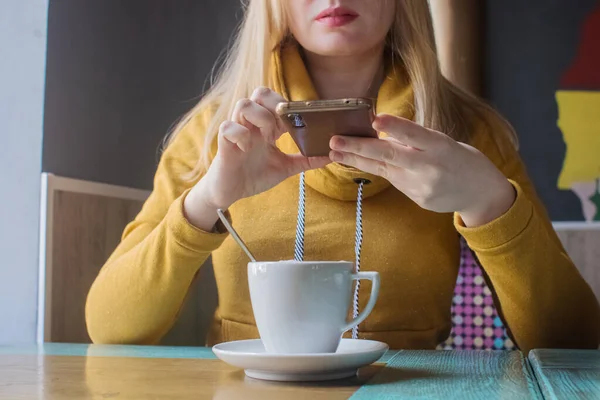 Blogger girl sits in a cafe and shoots a cappuccino foam on the smartphone. Work in social networks, video blogs and photo blogs. Concept for internet content creation and food photography.