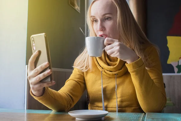 Blogcu kız bir kafede oturur ve akıllı telefondan kapuçino köpüğü atar. Sosyal ağlarda, video bloglarında ve fotoğraf bloglarında çalışıyorum. İnternet içeriği oluşturma ve gıda fotoğrafçılığı kavramı.
