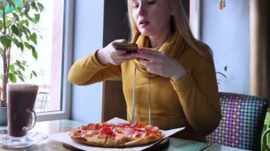 Blogcu bir kız bir kafede oturur ve akıllı telefondan pizza vurur. Sosyal ağlarda, video bloglarında ve fotoğraf bloglarında çalışıyorum. İnternet içeriği ve yiyecek fotoğrafçılığı oluşturma kavramı.