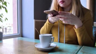 Blogcu kız bir kafede oturur ve akıllı telefondan kapuçino köpüğü atar. Sosyal ağlarda, video bloglarında ve fotoğraf bloglarında çalışıyorum. İnternet içeriği oluşturma ve gıda fotoğrafçılığı kavramı. 