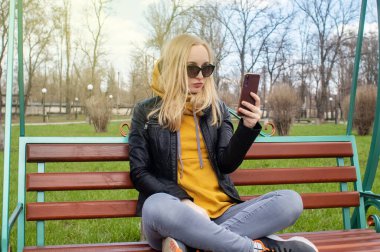 Blogcu kız parkta salıncakta oturuyor ve içerik yaratıyor. Sosyal ağlarda, video bloglarında ve fotoğraf bloglarında çalışıyorum. İnternet içeriği, eğitim ve çevrimiçi tanıtım kavramı