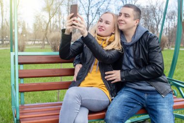 Karı koca parkta salıncakta sallanırken selfie çekiyorlar. Mutlu çift kendi fotoğraflarını çekiyor. Mutlu aile kavramı, poz vermek ve anı yakalamak..