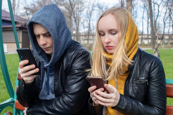 Man and a woman are both sitting on a date and sipping into their smartphones. Concept of social media addiction, getting out of reality and ignoring each other. Couple sitting on a swing in the park