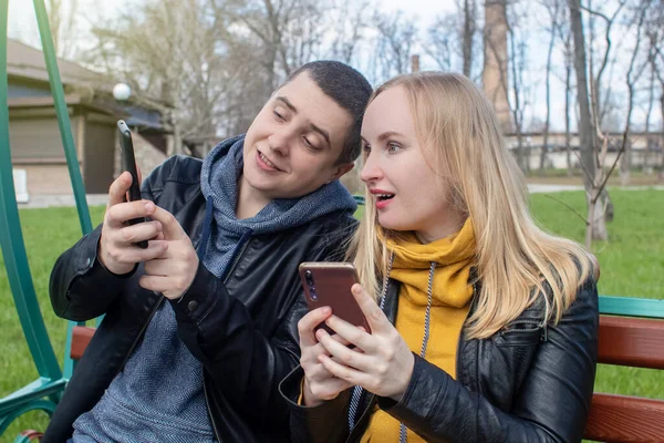 Man and a woman are both sitting on a date and sipping into their smartphones. Concept of social media addiction, getting out of reality and ignoring each other. Couple sitting on a swing in the park