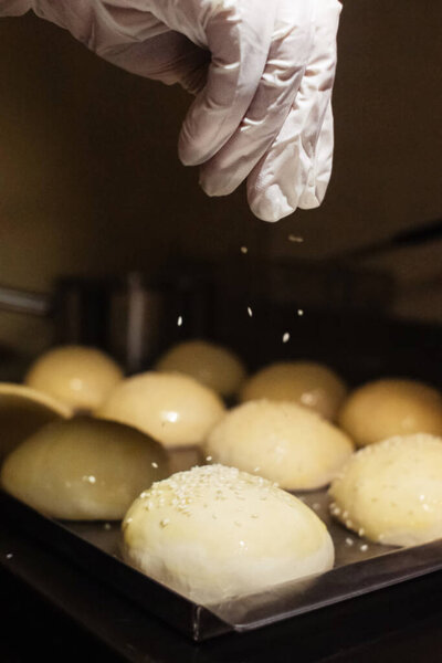 The baker sprinkles sesame seeds on the buns before placing them in the oven. Homemade baked goods and confectionery production. Home cooking concept. Vertical photo