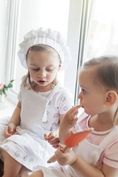 Children in the kitchen cook with their parents. Making homemade cake, pastries or cookies for the whole family. Happy childhood concept. Little twin sisters in kitchen aprons and chef hats