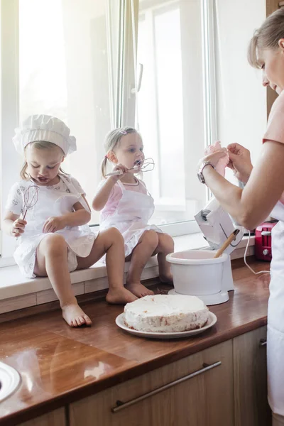 Children in the kitchen cook with their parents. Making homemade cake, pastries or cookies for the whole family. Happy childhood concept. Little twin sisters in kitchen aprons and chef hats