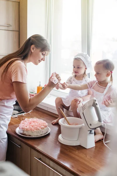 Children in the kitchen cook with their parents. Making homemade cake, pastries or cookies for the whole family. Happy childhood concept. Little twin sisters in kitchen aprons and chef hats