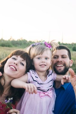 Anne, baba ve kızı sahada aile selfie 'si çekiyorlar. Doğada self-servis fotoğraf çekimi. Kızın saçında çiçekler var ve herkes çok mutlu ve gülümsüyor.