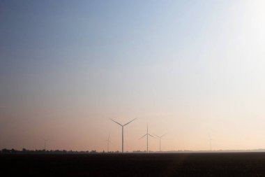 Sis altındaki yel değirmenleri, ayçiçeği tarlasının yanında. Sabah erkenden. Şiddetli bir rüzgar eserken pervanelerin hareketlerine bakın. Rüzgar türbini farklı mesafelere kurulur ve sisin içine gizlenir