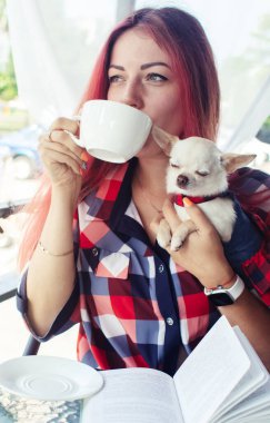 Kız bir kafede bir kitap okuyor. Yanındaki mini chihuahua, kahve ve kek. Dinlenme, rahatlama ve yaşam tarzı. Kızıl saçlı çekici bir kadın restoranda otururken hikaye okuyor..