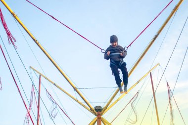 Çocuk bungee trambolinde zıplıyor. Sigortası ve esneyebilir lastikleri olan bir çocuk gökyüzüne asılı duruyor. Eğlence parkında mutlu çocukluk ve oyun kavramı.