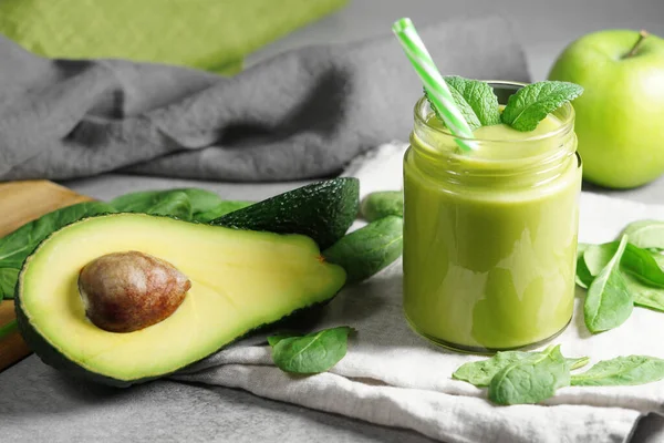 Avocado spinach and green apple smoothie healthy and refreshing tropical drink close-up view of a glass jar with mint leaves and sliced avocado fruit, apple and spinach leaves on grey kitchen board.