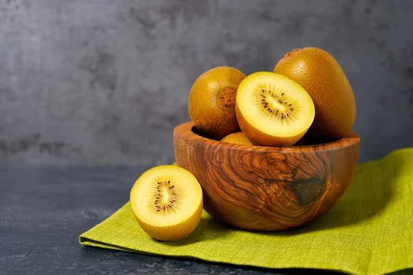 Kiwi gold yellow fruit in a wooden bowl with one sliced kiwi on grey background front view with copy space.