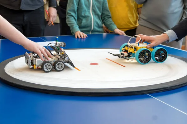 Yuvarlak beyaz yarışma sahasında (sumo) bir inşaat setinden toplanmış iki tekerlekli robotun yakın çekimi. Çocuklar ve yetişkinler mavi bir masanın etrafında durur, başlangıçtan önce mekanizmaları ayarlarlar..