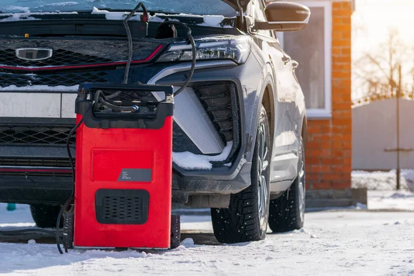 Profesyonel araba aküsü destekleyicisi kışın şarj oluyor. Kırmızı taşınabilir sıçrama başlatıcı ve akü şarj istasyonu kar üzerine park edilmiş modern bir arabaya bağlı..