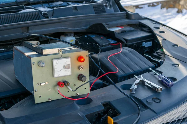Eski analog akü şarjı kışın kaputun altındaki bir araca bağlı. Klasik Analog Pil Şarj Aracı Kışın Araba Tamiri.
