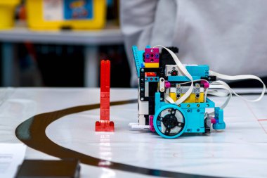 Mavi ve sarı bileşenleri olan özel yapım, modüler programlanabilir bir robot dikey kırmızı plastik bir engelin yanında bir yarışma alanı minderinin üzerine yerleştiriliyor. Alan çizgi-takip navigasyonu için eğri siyah çizgiler içerir.