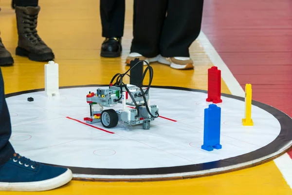 Robot tasnif ediliyor. Kırmızı çizgiler, eşmerkezli daireler ve renkli LEGO tuğla kuleleri (beyaz, kırmızı, mavi, sarı) ile büyük bir dairesel baskılı mat arenasına yerleştirilmiş gri bir LEGO robotu engeller veya hedefler.