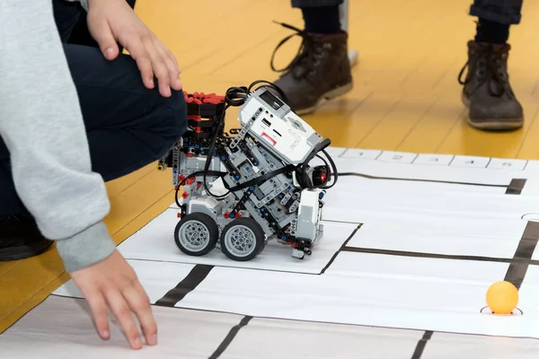 Spor salonunun zemininde çömelmiş, tekerlekleri, sensörleri, motorları ve kırmızı LEGO elementleri olan özel yapım LEGO robotunu yakından izleyen ve kontrol eden günlük kıyafetli bir çocuk..
