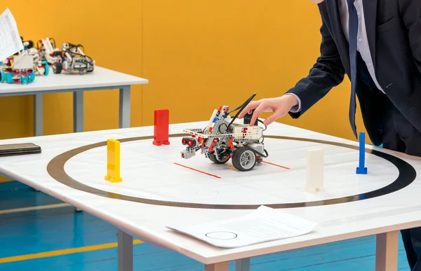 Resmi takım elbiseli bir öğrenci beyaz test masasındaki programlanabilir DIY robotunu ayarlamaya çalışıyor. Teknik dişliler, tekerlekler ve sensörlerden yapılmış robot renkli işaretlerin yer aldığı dairesel bir pist içinde yer alıyor. Robot sınıflandırılıyor