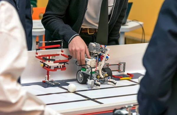 Okul üniformalı bir öğrenci, teknik bir atölye veya yarışma sırasında özel yapım bir robota ince ayar çekiyor. Robot modüler yapı taşları, vitesler, motorlar ve elektronik sensörlerden yapılmıştır.