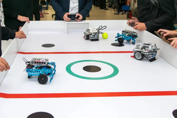 Kırmızı çizgili ve yeşil merkezli beyaz oyun alanında küçük, özel yapım robotları kontrol eden bir grup genç. Çevre. Robo futbol tarafından kontrol ediliyor.