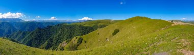 Great nature mountain range. Panorama perspective of caucasian mountain or volcano Elbrus with green fields, blue sky background. Elbrus landscape view - the highest peak of Russia and Europe