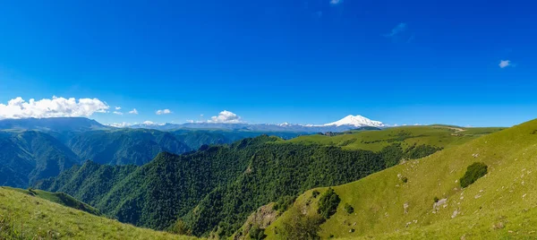Great nature mountain range. Panorama perspective of caucasian mountain or volcano Elbrus with green fields, blue sky background. Elbrus landscape view - the highest peak of Russia and Europe