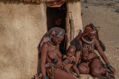 KORIXAS. NAMIBIA, 13 OCAK. 2020: Himba Kabilesi Kadınları Korixas, Namibya yakınlarındaki Himba köyündeki bir kulübenin önünde oturan bir çocukla birlikte