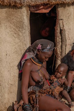 Himba kabilesinden Himba köyündeki bir kulübenin önünde çocuklu bir kadın oturuyor.