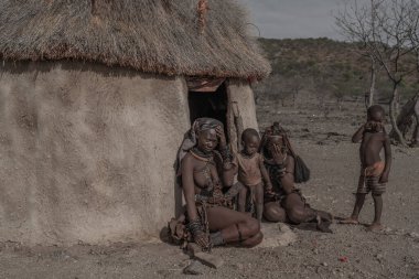 Himba köyündeki bir kulübenin önünde oturan iki çocuklu iki Himba kabilesi kadını.