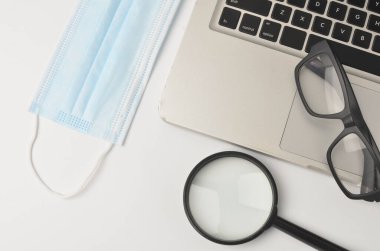 Beyaz ofis masası maskesi, büyüteç. Beyaz arka planda dizüstü bilgisayarı olan bir gözlük. Evden çalışmak ve covid-19 konsepti yeni normallik. Seçici odak.