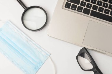 Beyaz ofis masası maskesi, büyüteç. Beyaz arka planda dizüstü bilgisayarı olan bir gözlük. Evden çalışmak ve covid-19 konsepti yeni normallik. Seçici odak.
