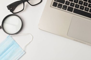 Beyaz ofis masası maskesi, büyüteç. Beyaz arka planda dizüstü bilgisayarı olan bir gözlük. Evden çalışmak ve covid-19 konsepti yeni normallik. Seçici odak.