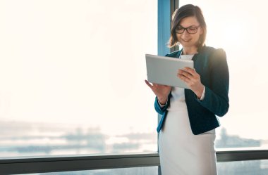 Şehre tepeden bakan bir ofis binasının penceresinin önünde dikilirken dijital tablet kullanan çekici bir iş kadını.
