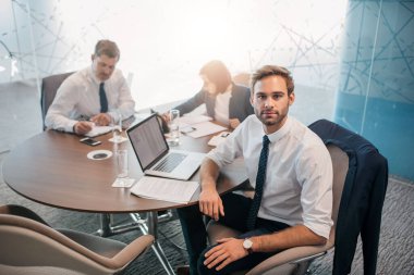 Genç bir iş adamının portresi arka planda iş arkadaşlarıyla bir masada otururken dizüstü bilgisayar kullanıyor.