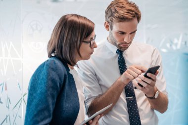 İki kendine güvenen genç iş adamı modern bir ofiste dururken cep telefonu ve dijital tablet üzerinden konuşuyorlar.