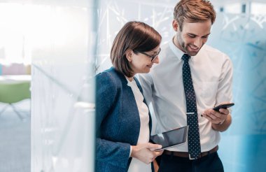 İki kendine güvenen genç iş adamı modern bir ofiste dururken cep telefonu ve dijital tablet üzerinden konuşuyorlar.