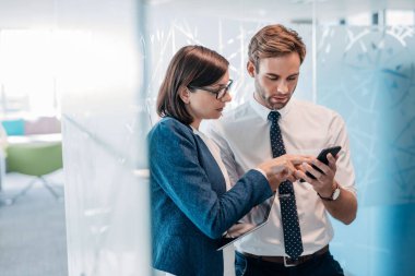 İki kendine güvenen genç iş adamı modern bir ofiste dururken cep telefonu ve dijital tablet üzerinden konuşuyorlar.