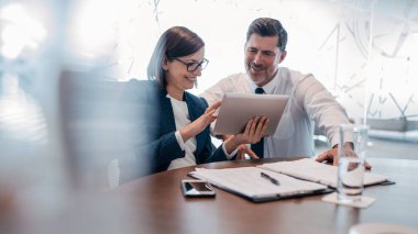 Modern bir ofiste masa başında çalışırken dijital tablet kullanan kendine güvenen iki iş adamı.