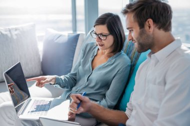 İki kendine güvenen iş adamı modern bir ofiste koltukta otururken dizüstü bilgisayar kullanıyor ve birlikte notlar okuyorlar.