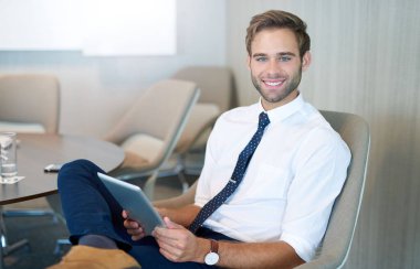 Modern genç bir işadamının portresi şık bir ofis alanında rahat bir şekilde otururken dijital tablet tutarken kameraya güvenle gülümsüyor.