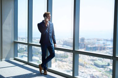 Modern bir ofisin en üst katındaki büyük pencerelerinin yanında duran şık genç bir işadamının tam uzunluktaki görüntüsü. Telefonuyla manzarayı seyrediyor.