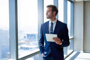 Modern ofiste elinde dijital tabletle büyük pencerelerden dışarı bakan şık takım elbiseli yakışıklı genç bir işadamı.