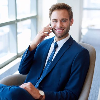 Ofisinde büyük camları olan yakışıklı genç bir işadamı kameraya gülümserken cep telefonuyla konuşuyor ve rahat bir modern sandalyede oturuyor.