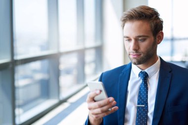 Geniş pencereli, parlak, modern bir ofiste duran şık genç işadamı cep telefonunun ekranına bakıyor.