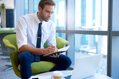 Genç bir işletmeci, günlüğünde notlar alırken bilgisayarına modern bir ofiste bakıyor.