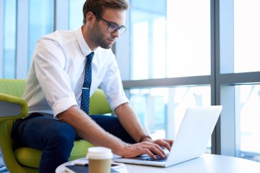 Genç iş adamı, modern bir ofiste kahve masasında oturmuş laptopunuzu daktilo ediyor.