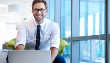 Modern ofis alanında genç bir işadamının portresi, önünde dizüstü bilgisayarıyla yazı yazıyor ve kameraya güvenle gülümsüyor.